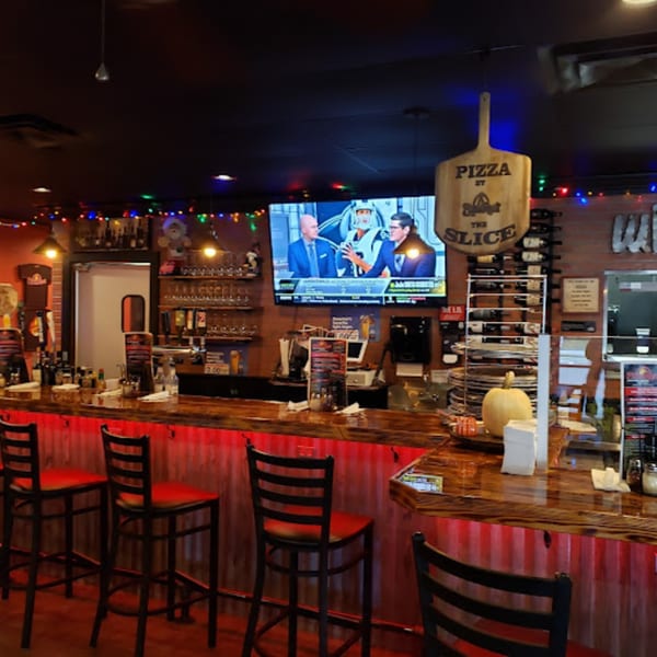 Pizza Rock's bar area with red bar stools, a TV, and a "Pizza by the Slice" sign.