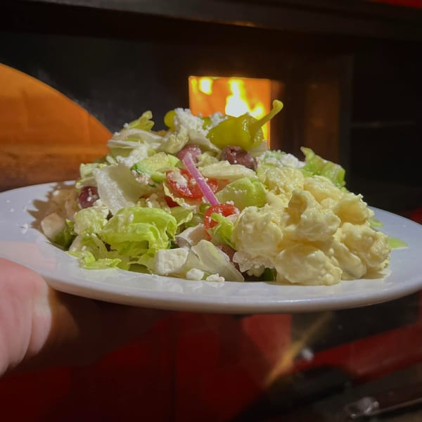 A fresh salad with lettuce, tomatoes, olives, feta, and macaroni salad, in front of a pizza oven.