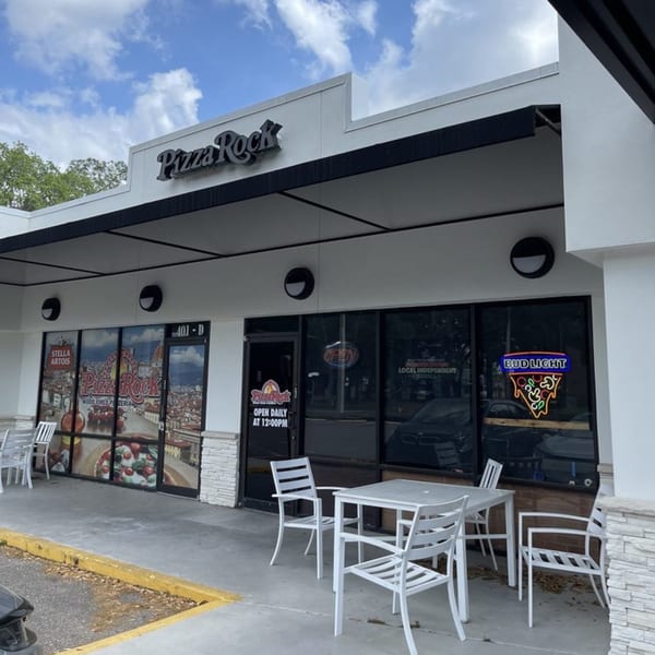 The exterior of Pizza Rock restaurant with outdoor seating, a black awning, and window decals.