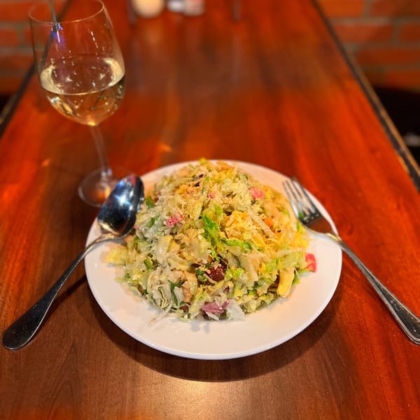 A large salad with shredded lettuce, cheese, and other toppings, next to a glass of white wine.